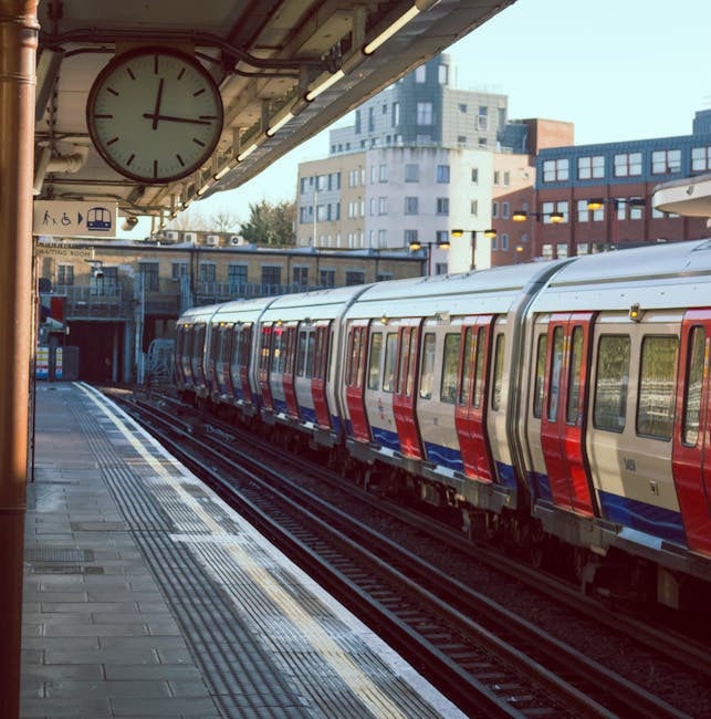 A modern passenger train with a sleek design and predominantly white exterior with purple and red accents is arriving at a suburban railway station platform on a clear day. The platform has tactile pa