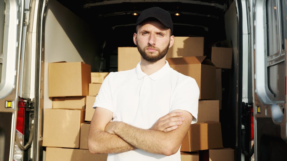 A man wearing casual clothing and a black cap is loading three cardboard boxes into the side of a white commercial van parked on a residential street in Chiswick. The van's rear and side doors are ope