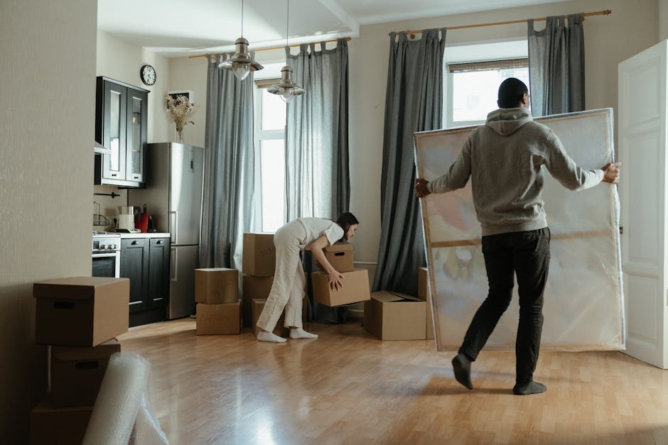 Inside a well-lit residential room during a home relocation, two individuals are engaged in packing and moving activities. The room features a light wooden floor, beige walls, and large windows with g