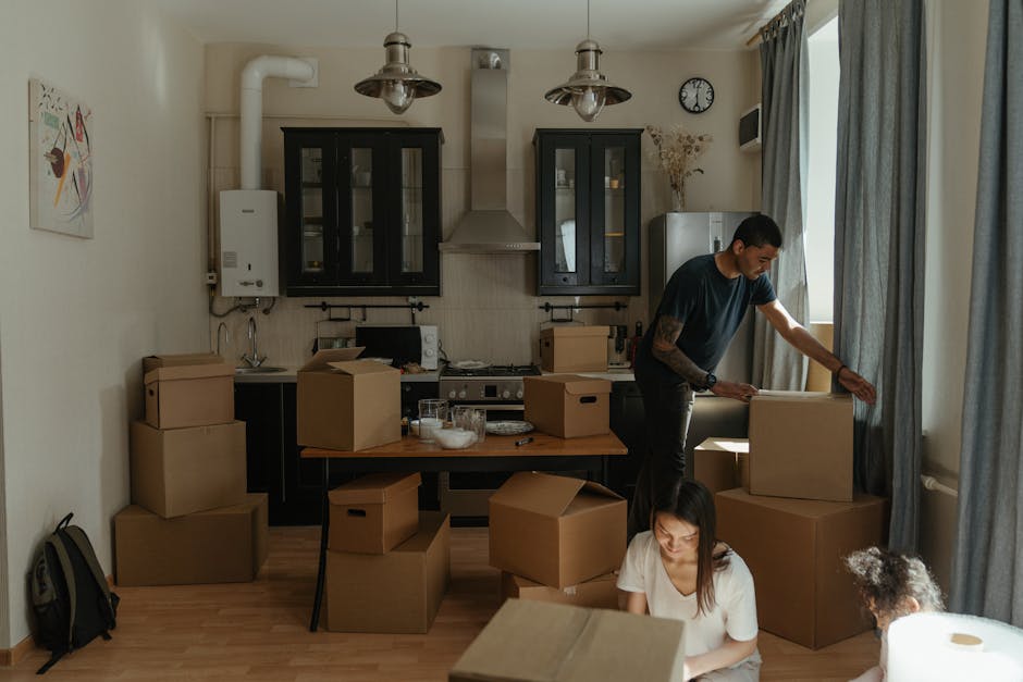 Inside a well-lit residential room during a home relocation, two individuals are engaged in packing and moving activities. The room features a light wooden floor, beige walls, and large windows with g